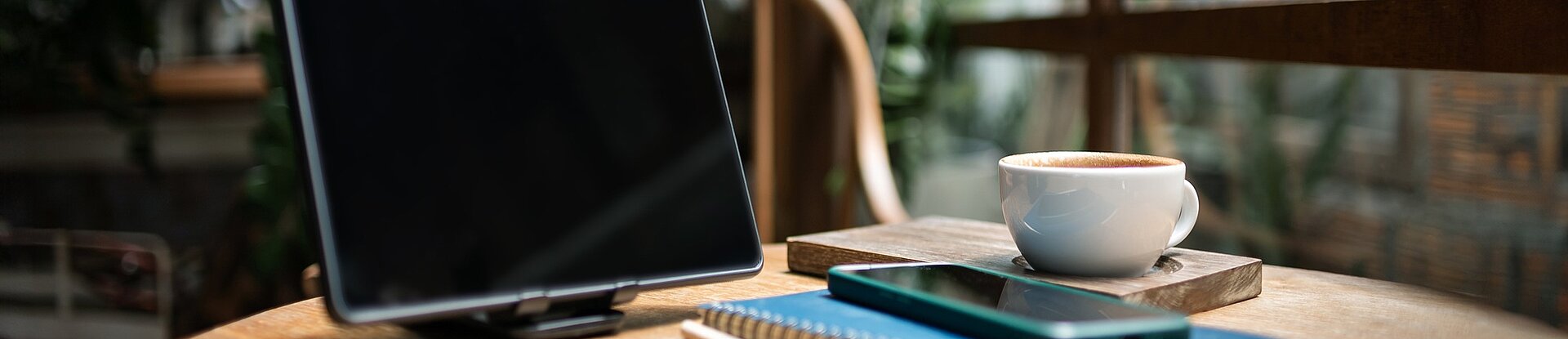 Ein Tablet steht neben einem blauen Notizblock, einem Handy und einer Tasse Kaffee auf einem runden Holztisch. Im Hintergrund sieht man einen Holzstuhl, eine Pflanze und eine Fensterfront.