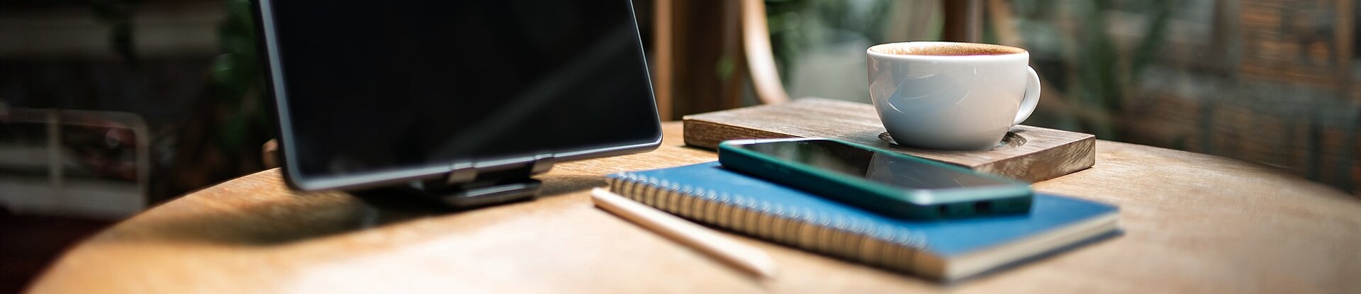 Tablet, Notizblock und Handy in einem Cafe Ein Tablet steht neben einem blauen Notizblock, einem Handy und einer Tasse Kaffee auf einem runden Holztisch. Im Hintergrund sieht man einen Holzstuhl, eine Pflanze und eine Fensterfront.