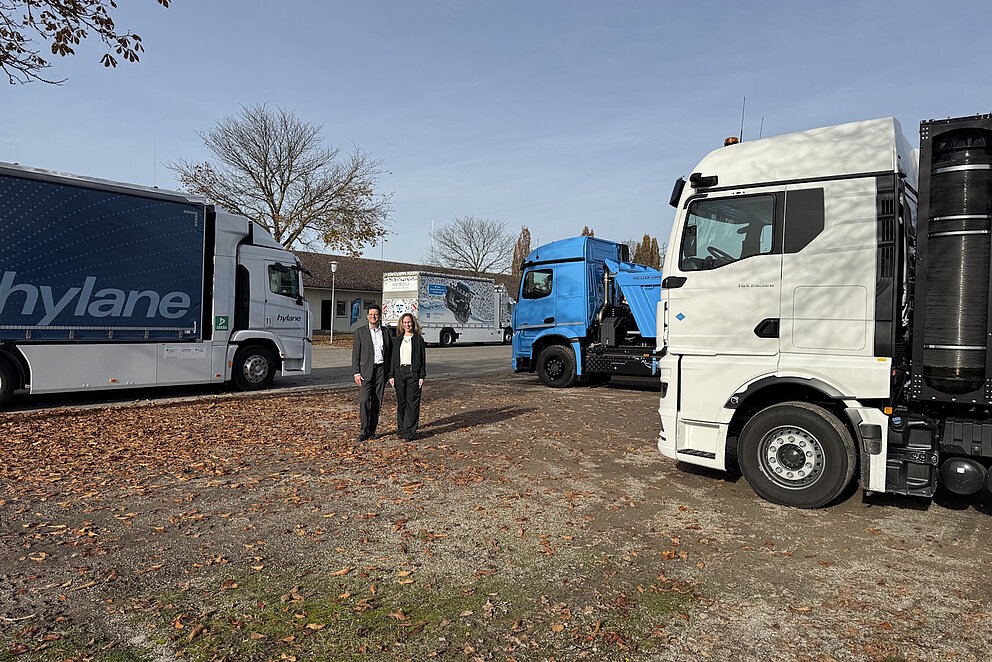 Vier Lkw stehen auf einer großen Fläche. In der Mitte stehen zwei Personen, die lächelnd in die Kamera blicken. 