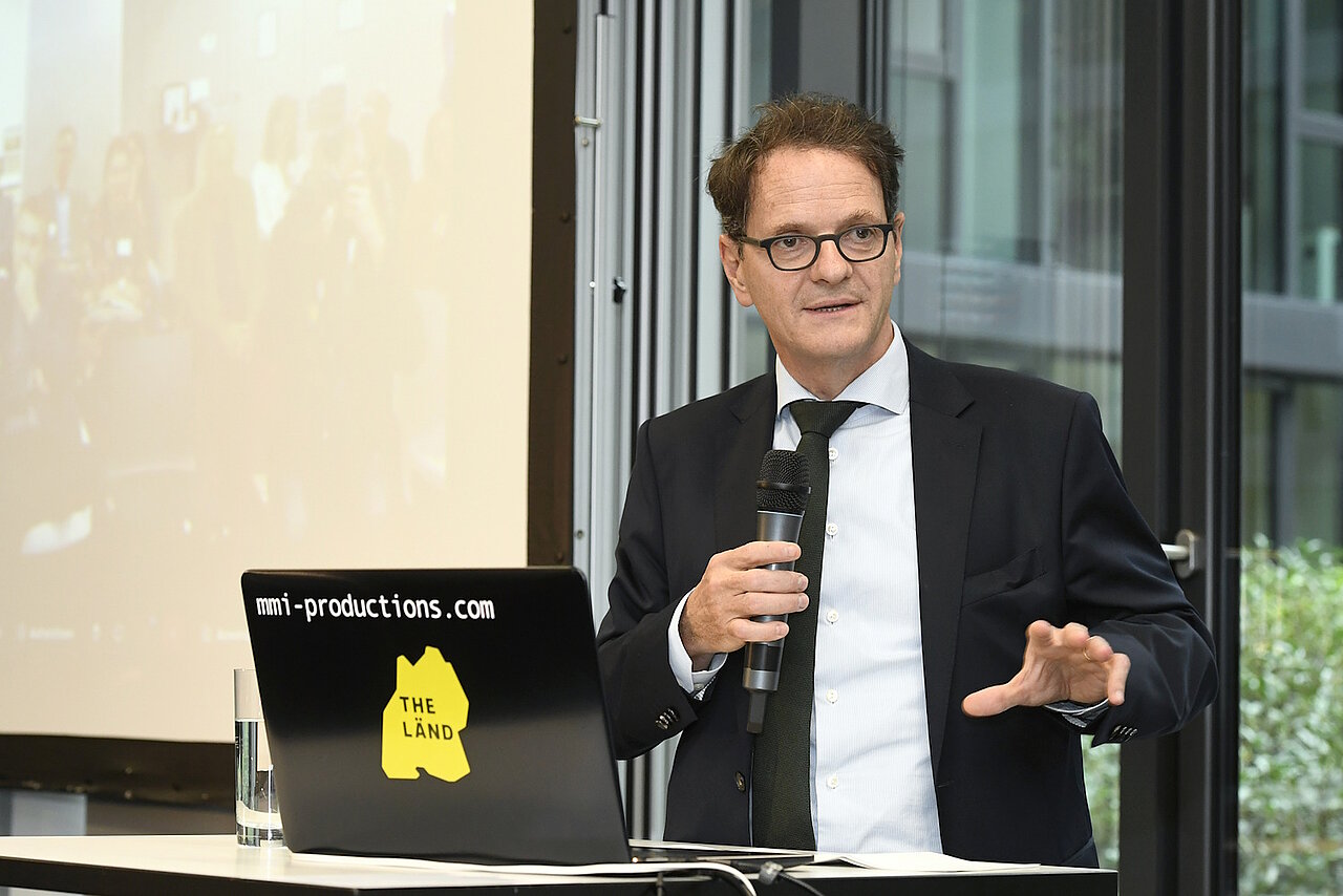 Ein Mann mit Brille hält einen Vortrag. In seiner Hand ist ein Mikrofon, neben ihm steht ein aufgeklappter Laptop.
