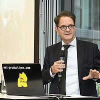 Ein Mann mit Brille hält einen Vortrag. In seiner Hand ist ein Mikrofon, neben ihm steht ein aufgeklappter Laptop.