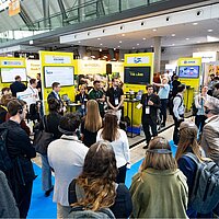 Eine Gruppe Menschen steht vor einem Messestand und hört einem Sprecher zu.