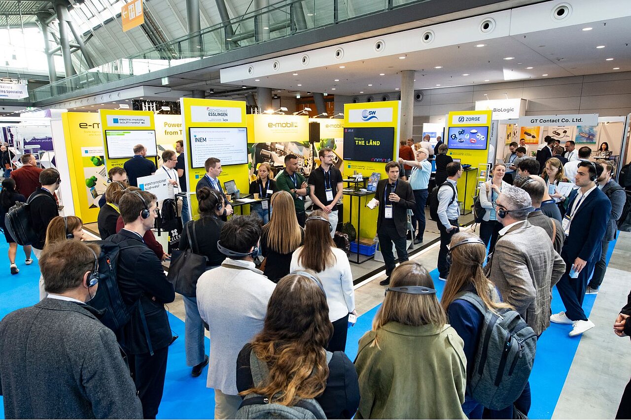 Eine Gruppe Menschen steht vor einem Messestand und hört einem Sprecher zu.