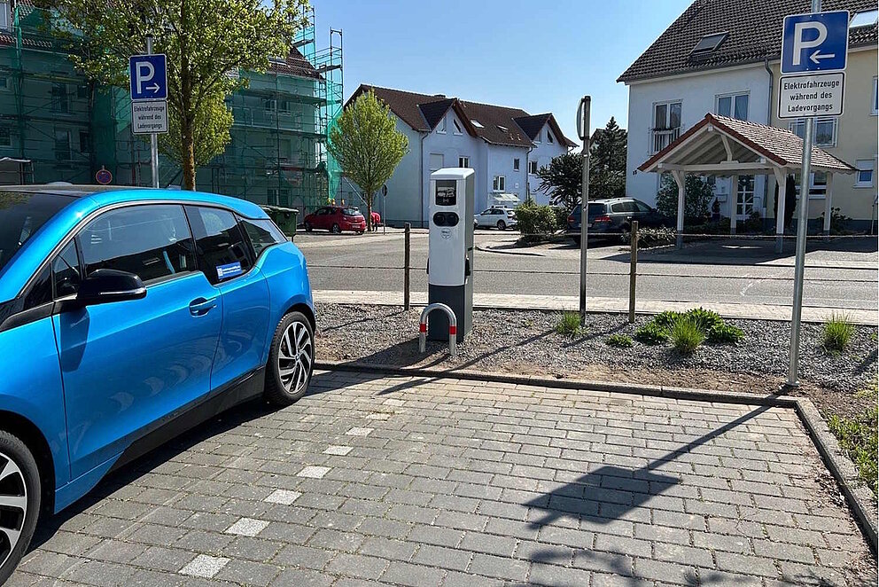 Ein blaues E-Auto steht auf einem Parkplatz neben einer Ladestation, dahinter sieht man ein Wohngebiet.