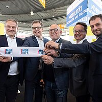 Fünf Personen halten das durchtrennte Band in den Händen, mit dem die i-Mobility eröffnet wurde. Die Personen lächeln dabei in die Kamera.