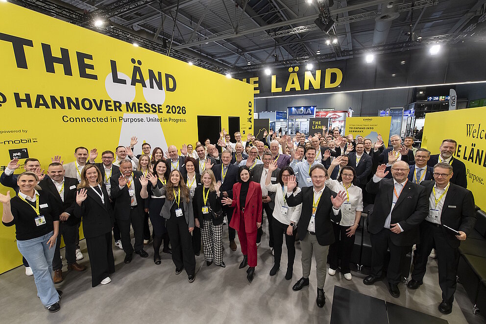 Winkende Menschen auf dem THE LÄND-Gemeinschaftsstand. 