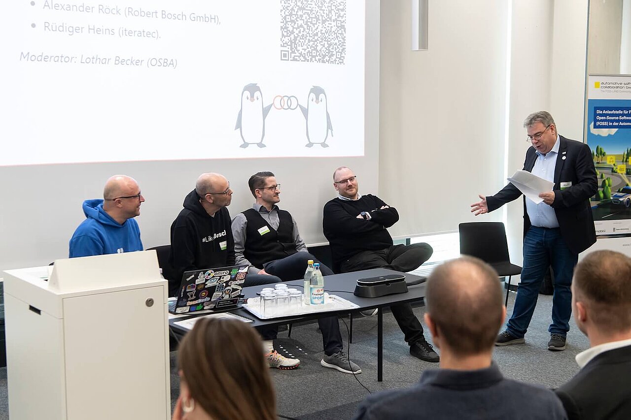 Die Speaker:innen der Veranstaltung OpenChain and Friends sitzen teilweise an ihren Plätzen. Ein Speaker steht und hält einen Vortrag.