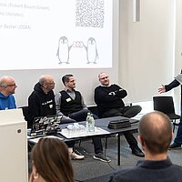 Die Speaker:innen der Veranstaltung OpenChain and Friends sitzen teilweise an ihren Plätzen. Ein Speaker steht und hält einen Vortrag.