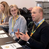 3 Menschen sitzen an einem Tisch. Ein Mann redet, die anderen hören zu.,