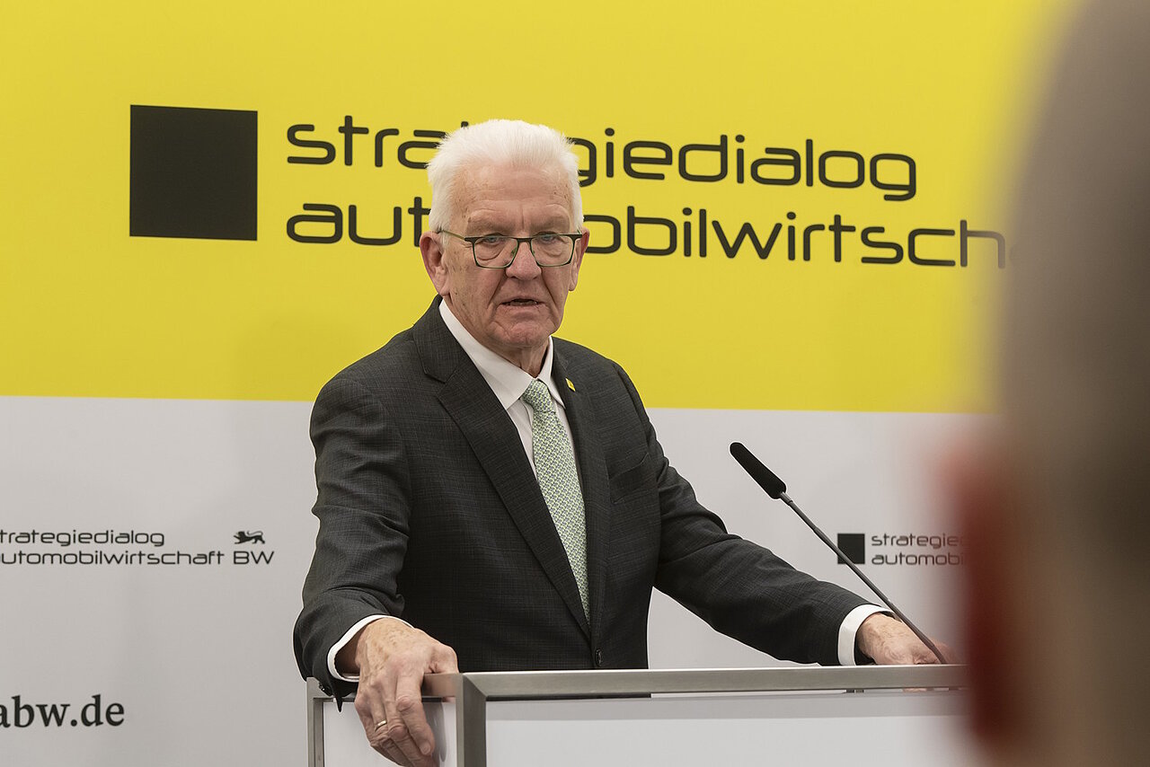 Ministerpräsident Winfried Kretschmann bei einer Pressekonferenz vor einer gelben des Strategiedialog Automobilwirtschaft BW..