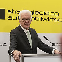 Ministerpräsident Winfried Kretschmann bei einer Pressekonferenz vor einer gelben des Strategiedialog Automobilwirtschaft BW..