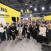 Eine große Gruppe an Personen steht im Halbkreis auf einem Messestand und winkt in die Kamera.