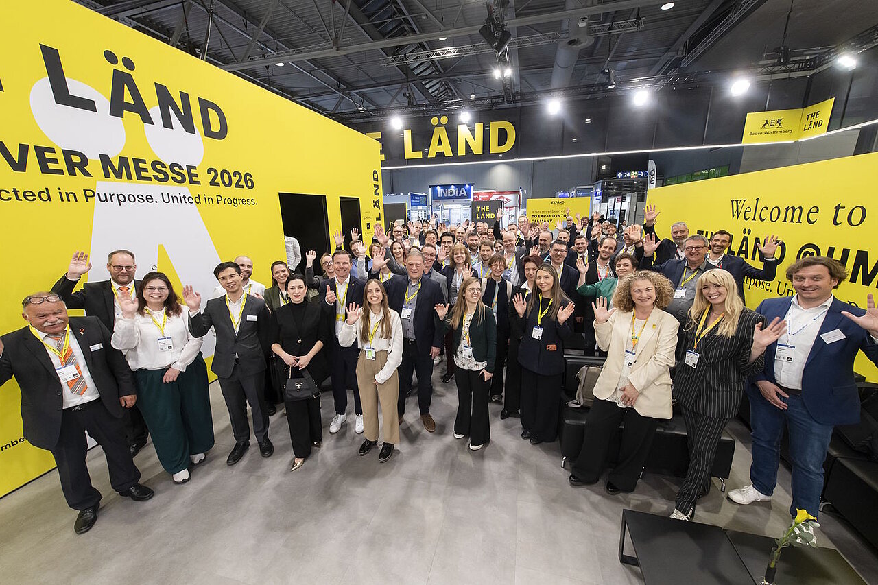 Eine große Gruppe an Personen steht im Halbkreis auf einem Messestand und winkt in die Kamera.