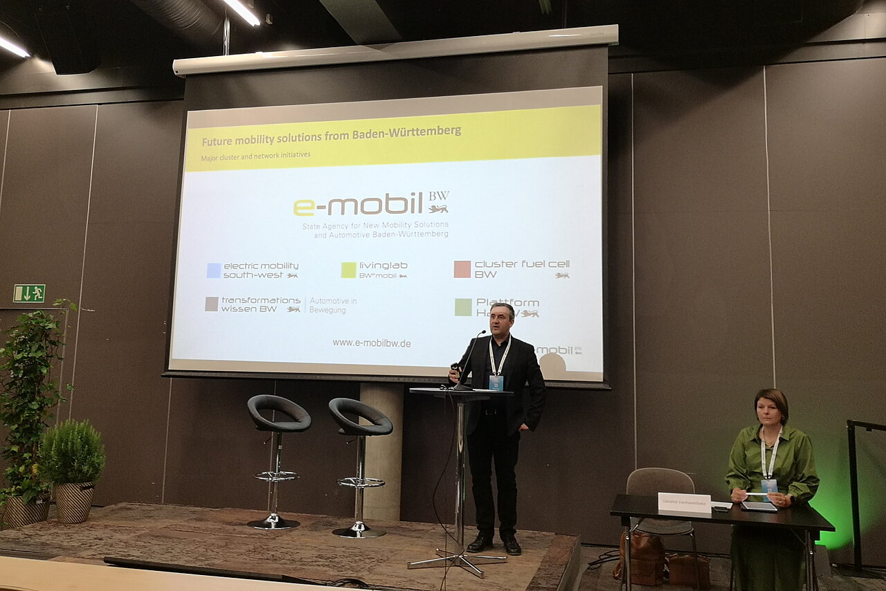 Dr. Wolfgang Fischer hält einen Vortrag und im Hintergrund ist eine PowerPoint Folie mit e-mobil BW Logos zu sehen. 