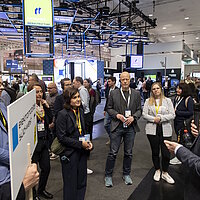 Mehrere Personen stehen im Halbkreis um einen Messestand, ein Aussteller erklärt etwas.