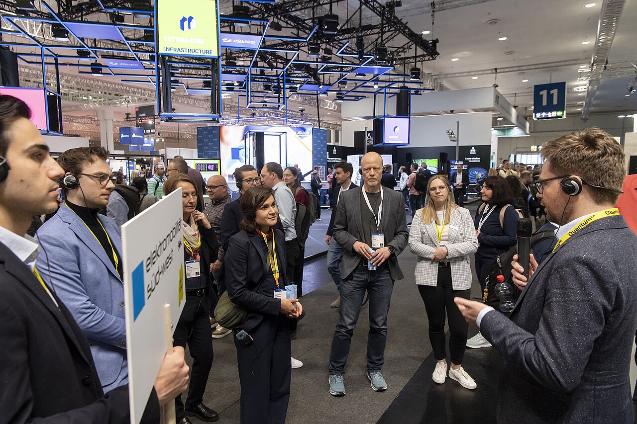 Mehrere Personen stehen im Halbkreis um einen Messestand, ein Aussteller erklärt etwas.