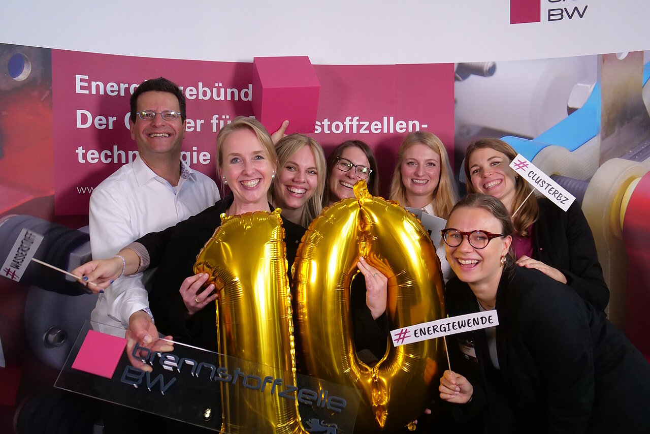 Gruppenmfoto des Teams Wasserstofftechnologien der e-mobil BW, einige halten Schilder in der Hand mit der Aufschrift "#Wasserstoff" und die Person im Vordergrund hält eine "10" aus zwei goldenen Ballons in der Hand.