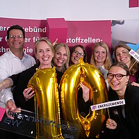 Gruppenmfoto des Teams Wasserstofftechnologien der e-mobil BW, einige halten Schilder in der Hand mit der Aufschrift "#Wasserstoff" und die Person im Vordergrund hält eine "10" aus zwei goldenen Ballons in der Hand.