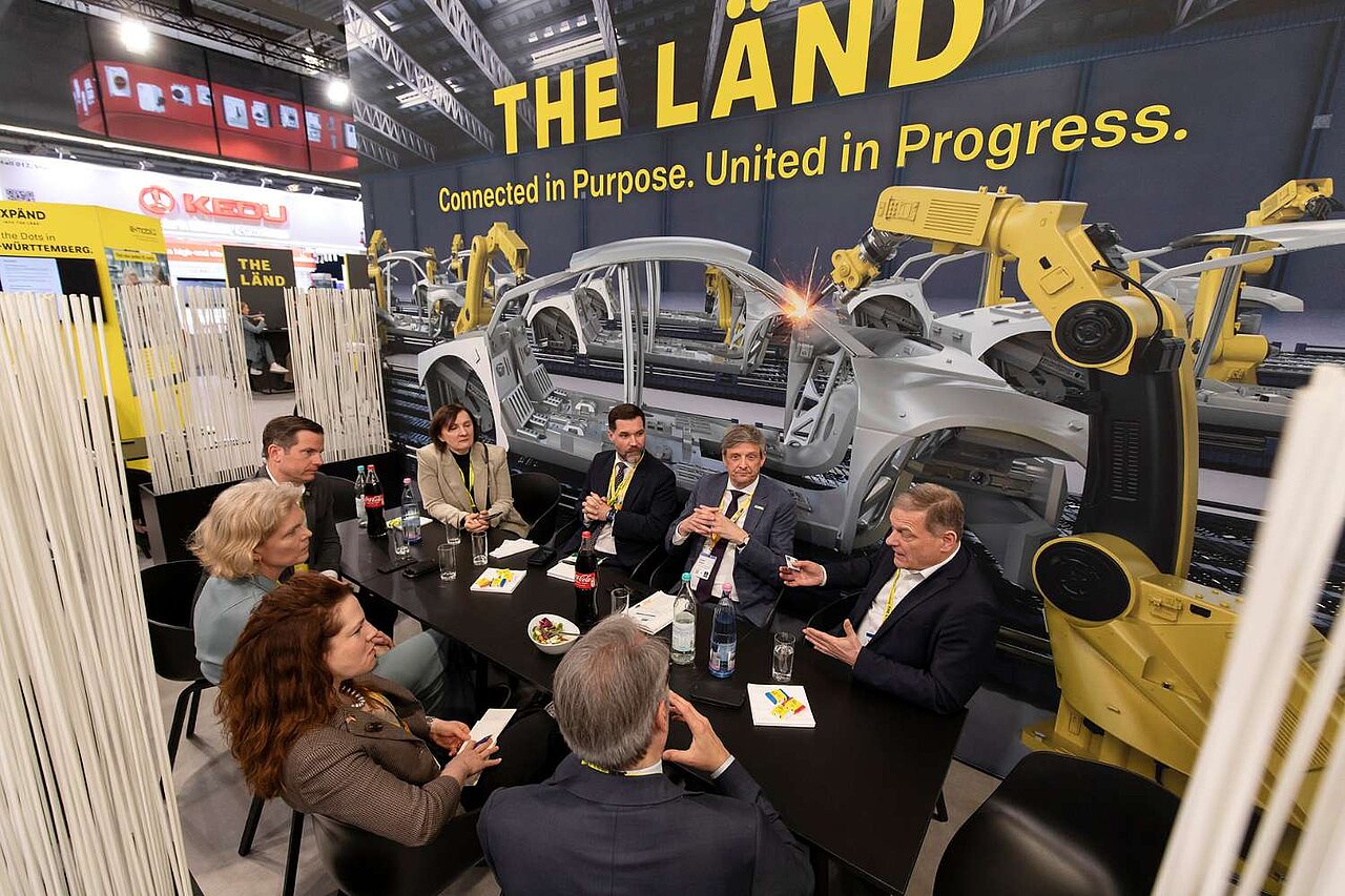 Eine Gruppe an Menschen sitzen im Kreis um einen Tisch auf einer Messe und unterhalten sich.