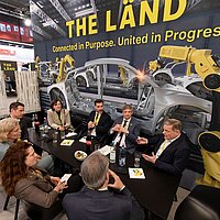 Eine Gruppe an Menschen sitzen im Kreis um einen Tisch auf einer Messe und unterhalten sich.