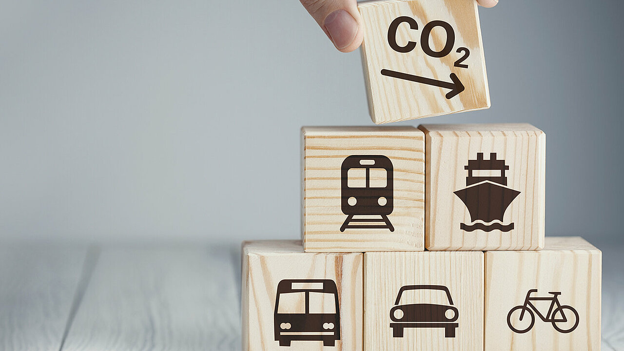 Fünf aufeinandergestapelte Würfel aus Holz mit Symbolen, die den Verlehrssektor repräsentieren (Bus, PKW, Fahrrad, Zug, Schiff) und ein Würfel mit einem Symbol für sinkende CO2-Emissionen
