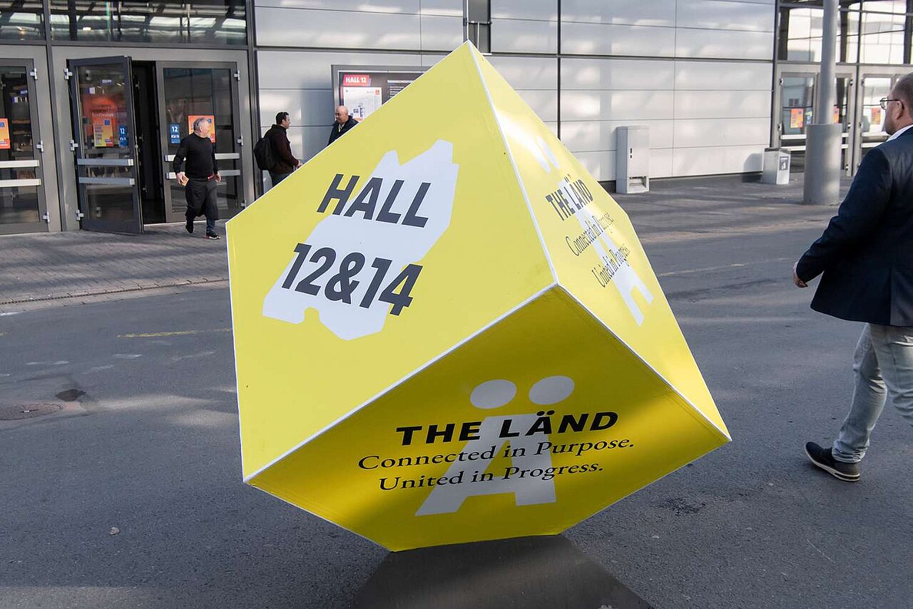 Ein gelber Quader mit der Aufschrift "Halle 12 & 14 THE LÄND" steht auf dem Boden vor der Messe Hannover.