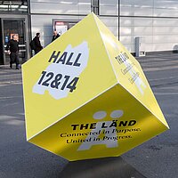 Ein gelber Quader mit der Aufschrift "Halle 12 & 14 THE LÄND" steht auf dem Boden vor der Messe Hannover.