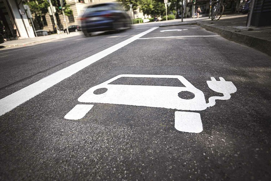 Auf der Straße ist im Vordergrund ein weißes Auto-Symbol mit Stecker zu sehen. Im Hintergrund fährt ein Auto vorbei.