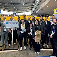 Eine Personengruppe posiert für ein Gruppenfoto auf einem gelben Messestand der e-mobil BW.