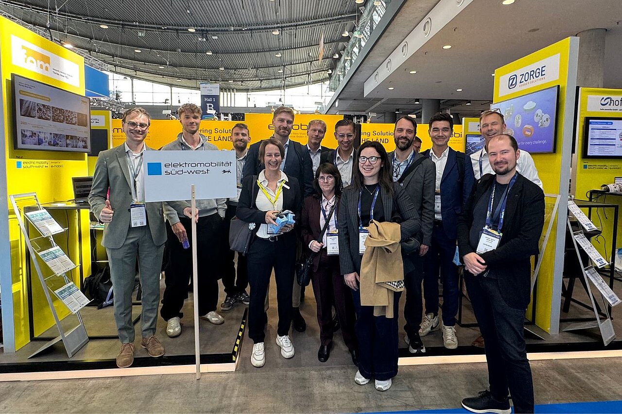 Eine Personengruppe posiert für ein Gruppenfoto auf einem gelben Messestand der e-mobil BW.