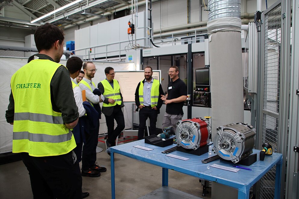 6 Männer stehen in einer Maschinenhalle um einen Tisch mit drei E-Motoren-Bauteilen