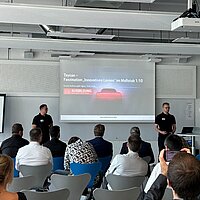 Zwei Männer in schwarzer Kleidung präsentieren vor einem Publikum eine PowerPoint-Folie mit dem Titel „Taycan – Faszination ‚Innovatives Lernen‘ im Maßstab 1:10“ in einem modernen Seminarraum. Im Raum sitzen mehrere Personen, einige machen Fotos oder schauen auf ihr Handy. 