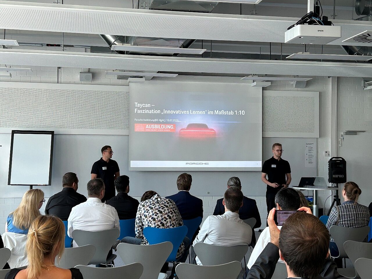 Zwei Männer in schwarzer Kleidung präsentieren vor einem Publikum eine PowerPoint-Folie mit dem Titel „Taycan – Faszination ‚Innovatives Lernen‘ im Maßstab 1:10“ in einem modernen Seminarraum. Im Raum sitzen mehrere Personen, einige machen Fotos oder schauen auf ihr Handy. 