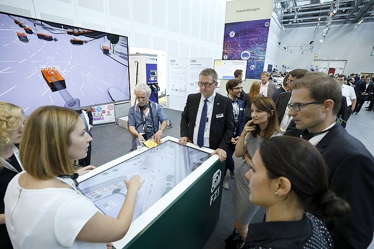 Besuchende einer Messe stehen vor einem digitalen Informationstisch und lassen sich die Themenfelder des Strategiedialog Automobilwirtschaft BW erklären.