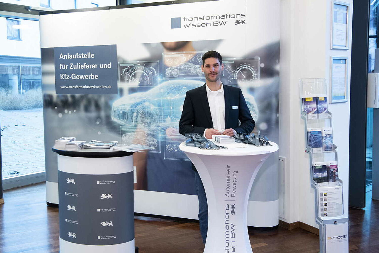 Ein Informationsstand der Landeslotsenstelle Transformationswissen BW mit Tisch und Aufsteller, dahinter steht Tim Siegel der e-mobil BW.