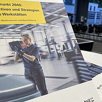 Es ist das Studiencover der Studie "Servicemarkt 2040: Perspektiven und Strategien für freie Kfz-Werkstätten" zu sehen, rechts im Hintergrund Ausblick in einen Saal mit Sitzplätzen.
