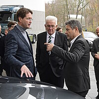Baden-Württembergs Ministerpräsident Winfried Kretschmann steht mit zwei Personen vor einem Mercedes Pkw und unterhält sich.