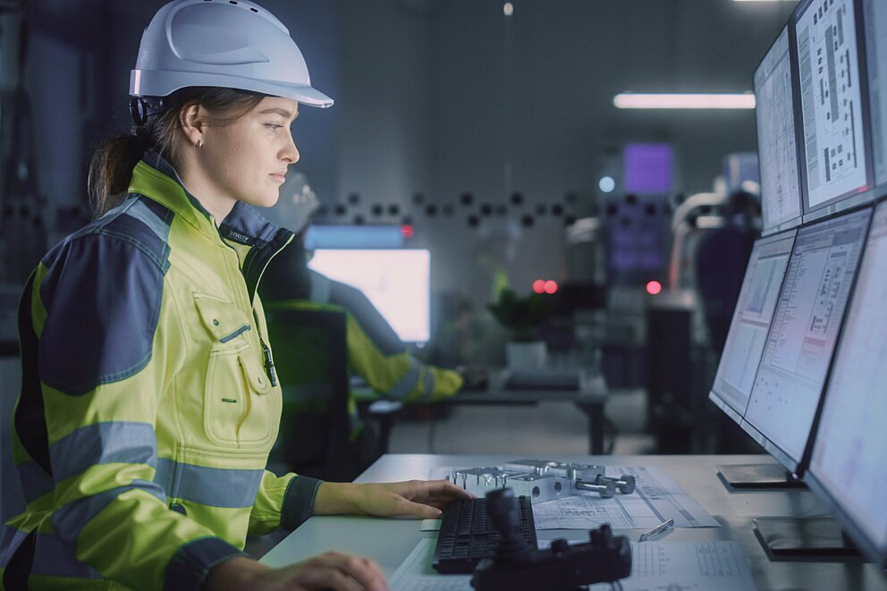 Seitliche Aufnahme einer jungen Frau mit Schutzhelm und Arbeitskleidung. Sie blickt konzentriert auf mehrere große Bildschirme mit technischen Daten.