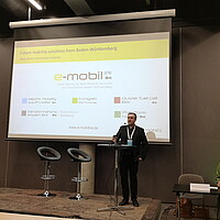 Dr. Wolfgang Fischer hält einen Vortrag und im Hintergrund ist eine PowerPoint Folie mit e-mobil BW Logos zu sehen. 