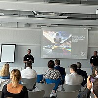 Ein Mann in schwarzer Kleidung steht vor einer Leinwand in einem Seminarraum.