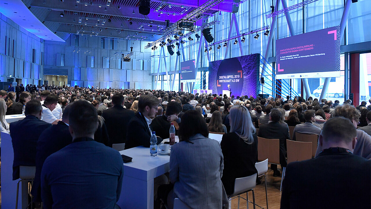 Das Foto des Digitalgipfel BW 2019 zeigt einen Konferenzsaal mit Bühne und vielen Besucher:innen, das Licht ist bläulich gefärbt.