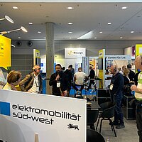 Eine Personengruppe steht auf einem gelben Messestand der e-mobil BW zusammen.