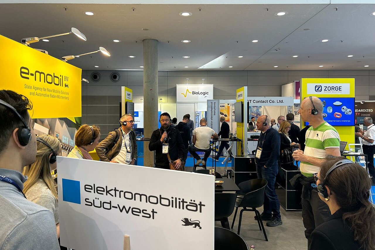 Eine Personengruppe steht auf einem gelben Messestand der e-mobil BW zusammen.