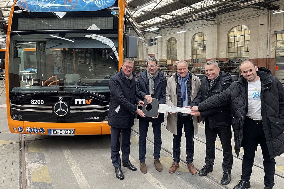 Eine gruppe Männer steht vor Wasserstoffbus in einer Halle und halten einen überdimensionierten Schlüssel in den Händen.