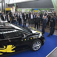 Messestand der e-mobil BW. Im Vordergrund ein elektrisches Auto, im Hintergrund Messebesucher:innen. 