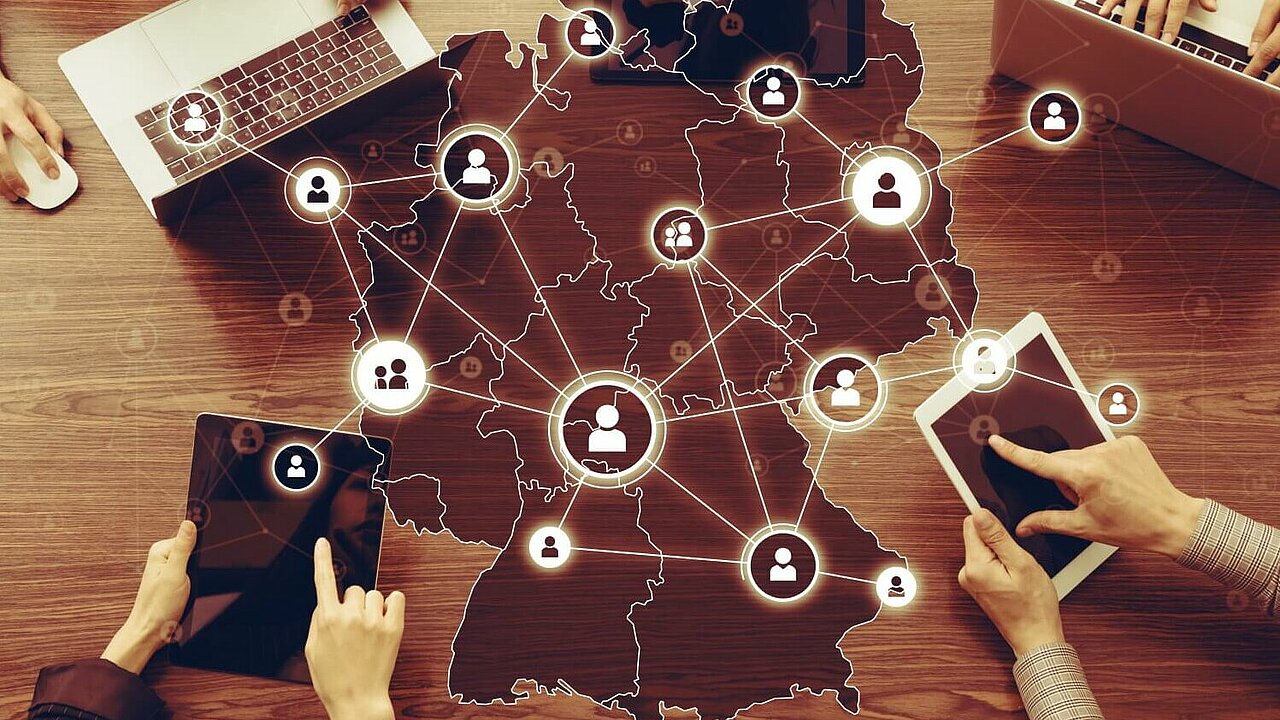 ünf Personen sitzen um einen Tisch, jeweils mit unterschiedlichen elektronischen Geräten vor sich, wie Laptops oder Tablets. In der Mitte des Tisches ist die Umrisskarte von Deutschland zu sehen. Linien verbinden die Geräte mit der Karte. Zwischen den Linien sind zahlreiche Icons von stilisierten Personen abgebildet, die Kontakte oder Netzwerke symbolisieren.