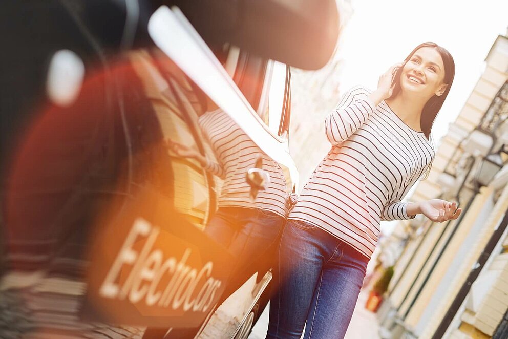 Eine junge Frau telefoniert mit einem Handy und steht dabei neben einem Elektroauto. 