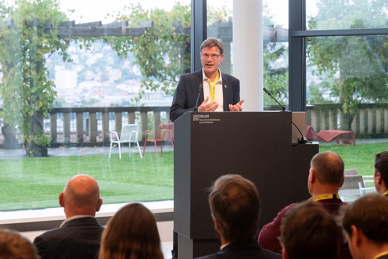 Christoph Erdmenger vom Verkehrsministerium Baden-Württemberg steht bei einer Veranstaltung auf einem Podium und spricht zum Publikum.
