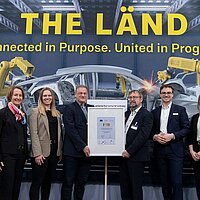 Sieben Personen stehen auf einer Messe vor einem Aufsteller, darunter Mitarbeitende des Clusters Elektromobilität Süd-West sowie der Geschäftsführer der e-mobil BW, Franz Loogen. In der Mitte wird die Auszeichnung ‚Gold Label‘ an das Cluster übergeben.