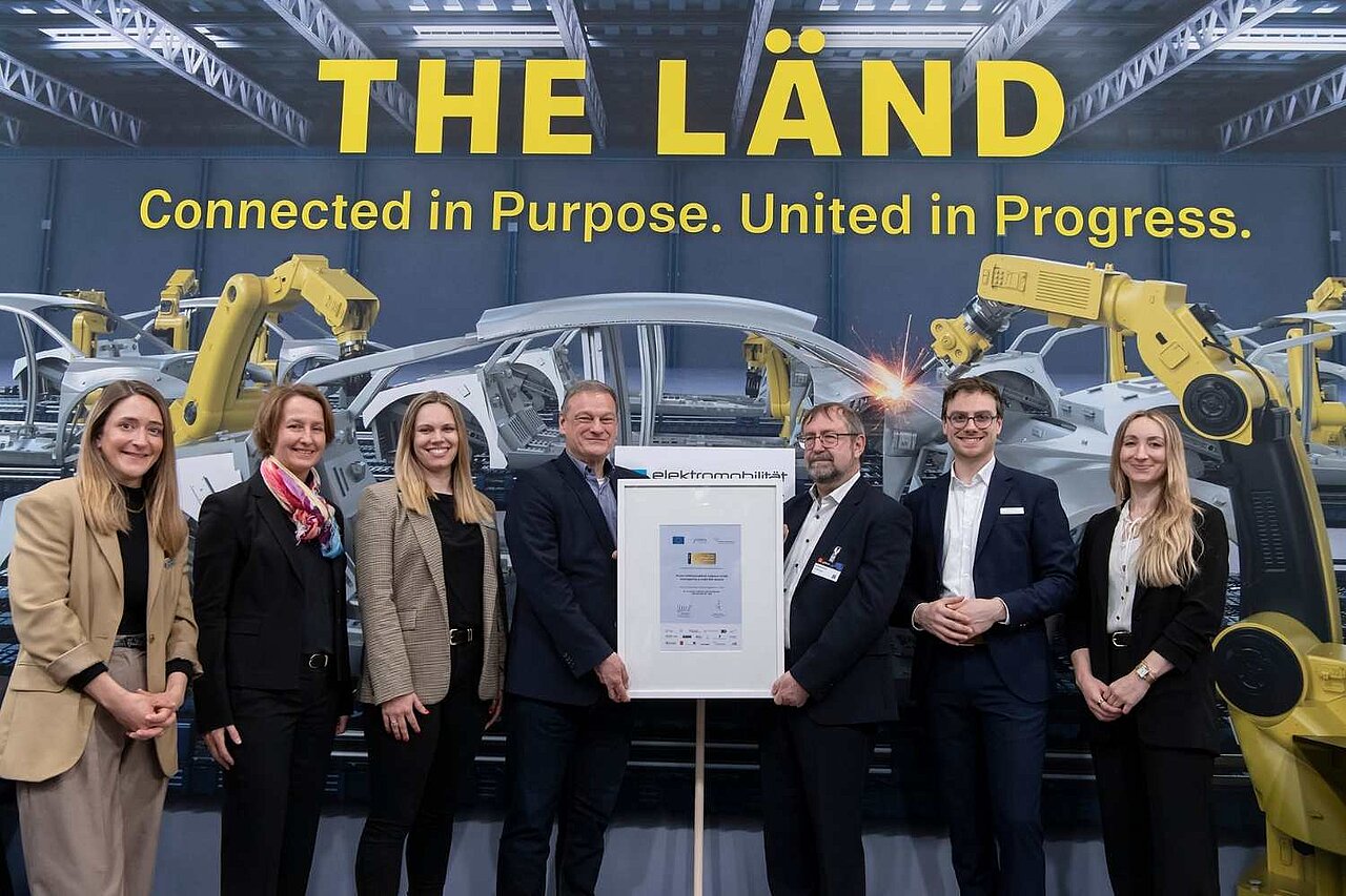 Sieben Personen stehen auf einer Messe vor einem Aufsteller, darunter Mitarbeitende des Clusters Elektromobilität Süd-West sowie der Geschäftsführer der e-mobil BW, Franz Loogen. In der Mitte wird die Auszeichnung ‚Gold Label‘ an das Cluster übergeben.